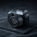 A miniature black camera-shaped device with a textured lens cover, white camera lens markings, and a small gray rectangular window on top, shown on a plain white background.